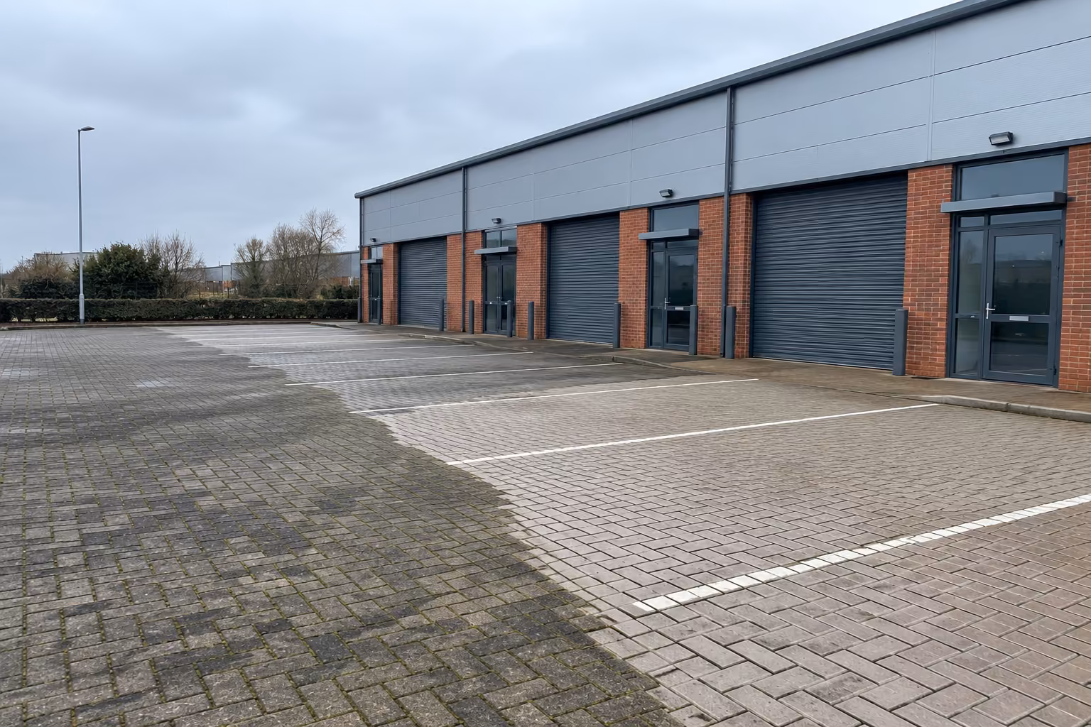Block-paved car park outside a row of brick and steel trade units, half pressure washed, showing the clean paving next to the dirty section